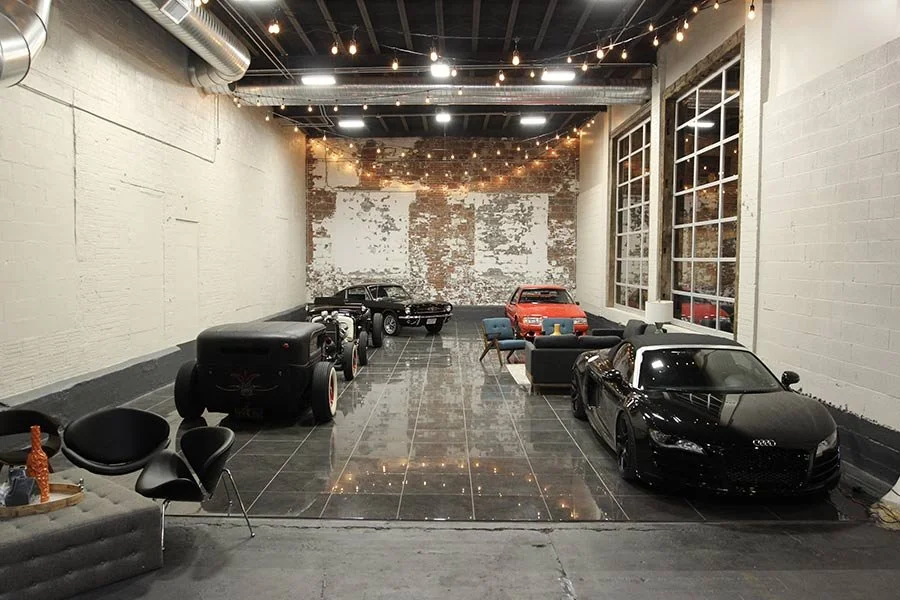 The Fuel House garage interior — member stalls with classic cars, hot rod, and Audi R8 under string lights
